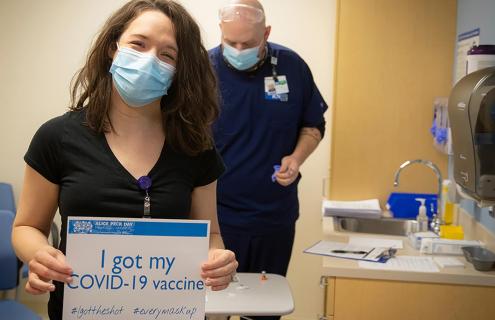 A woman, wearing a mask, holding a sign that says "I got my COVID-19 vaccine"